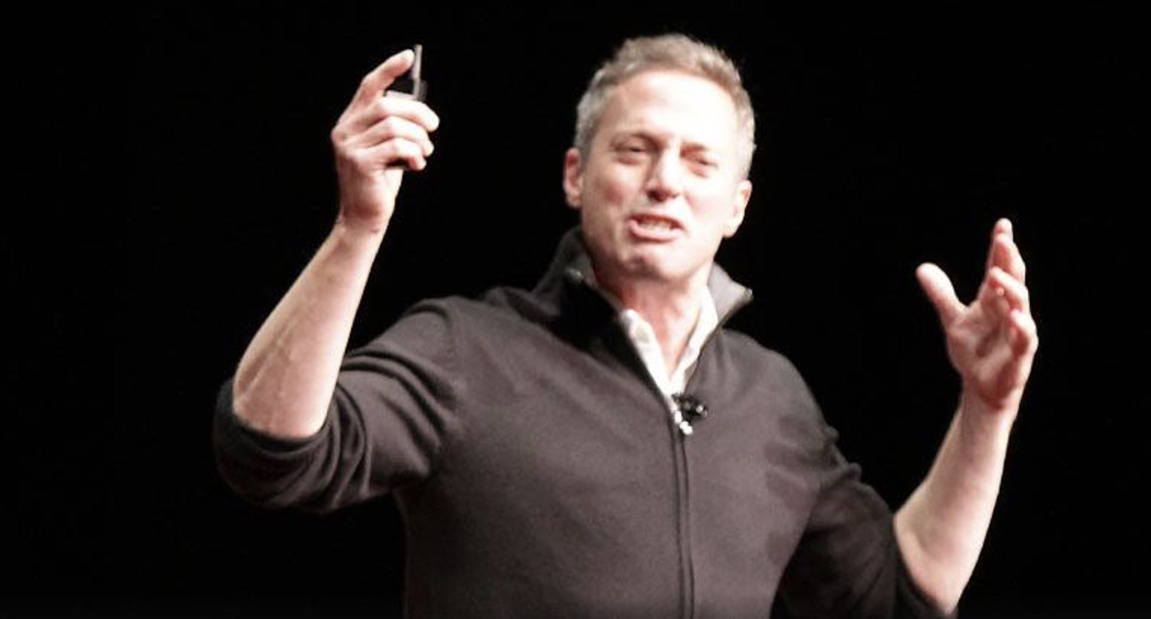 Light-skinned man with grey hair stands against a backdrop and makes a pointed gesture with his hand.