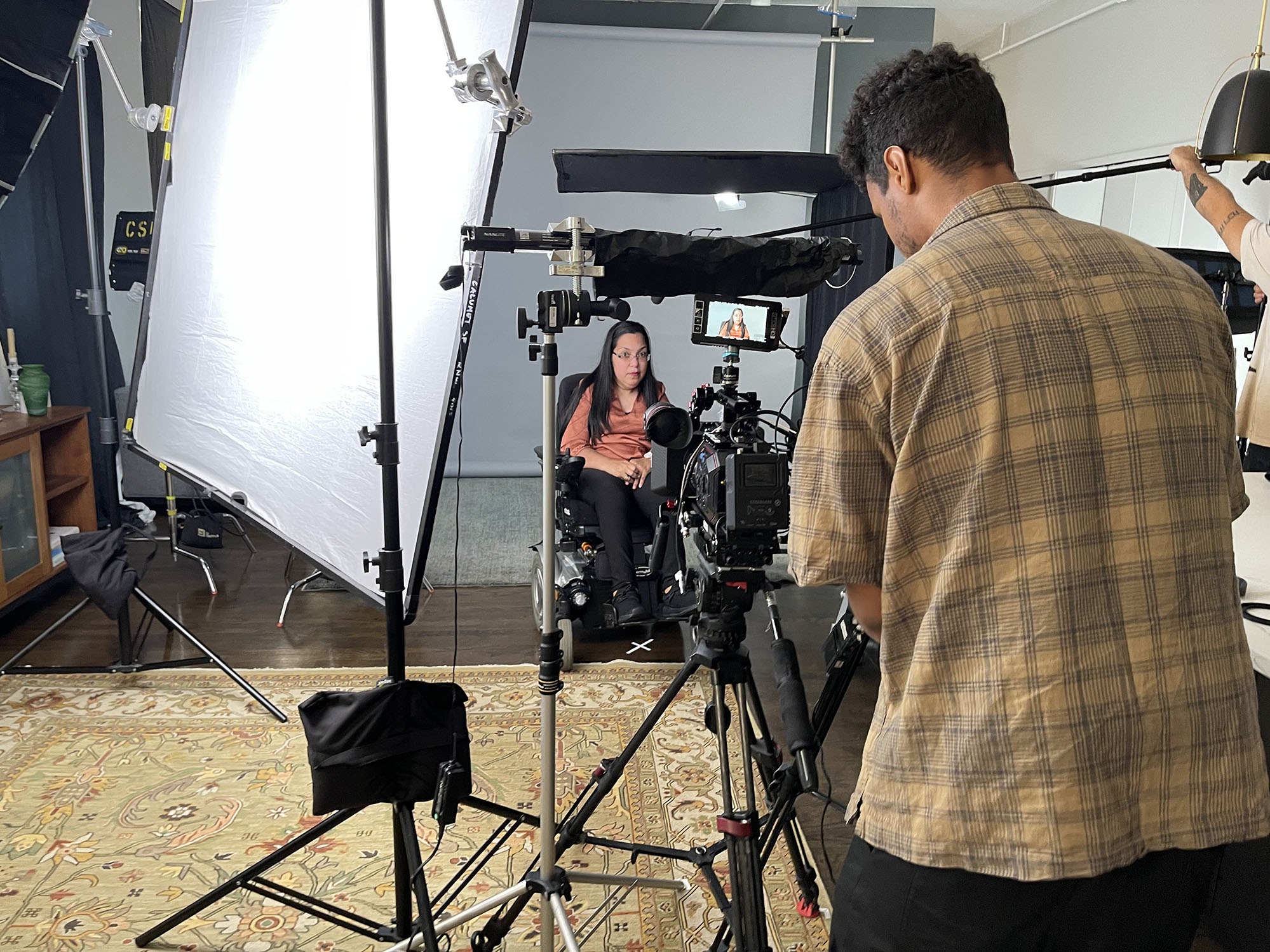 Medium-skinned woman in a wheelchair, on set of a movie. Medium-skinned man in foreground looking through camera lens.