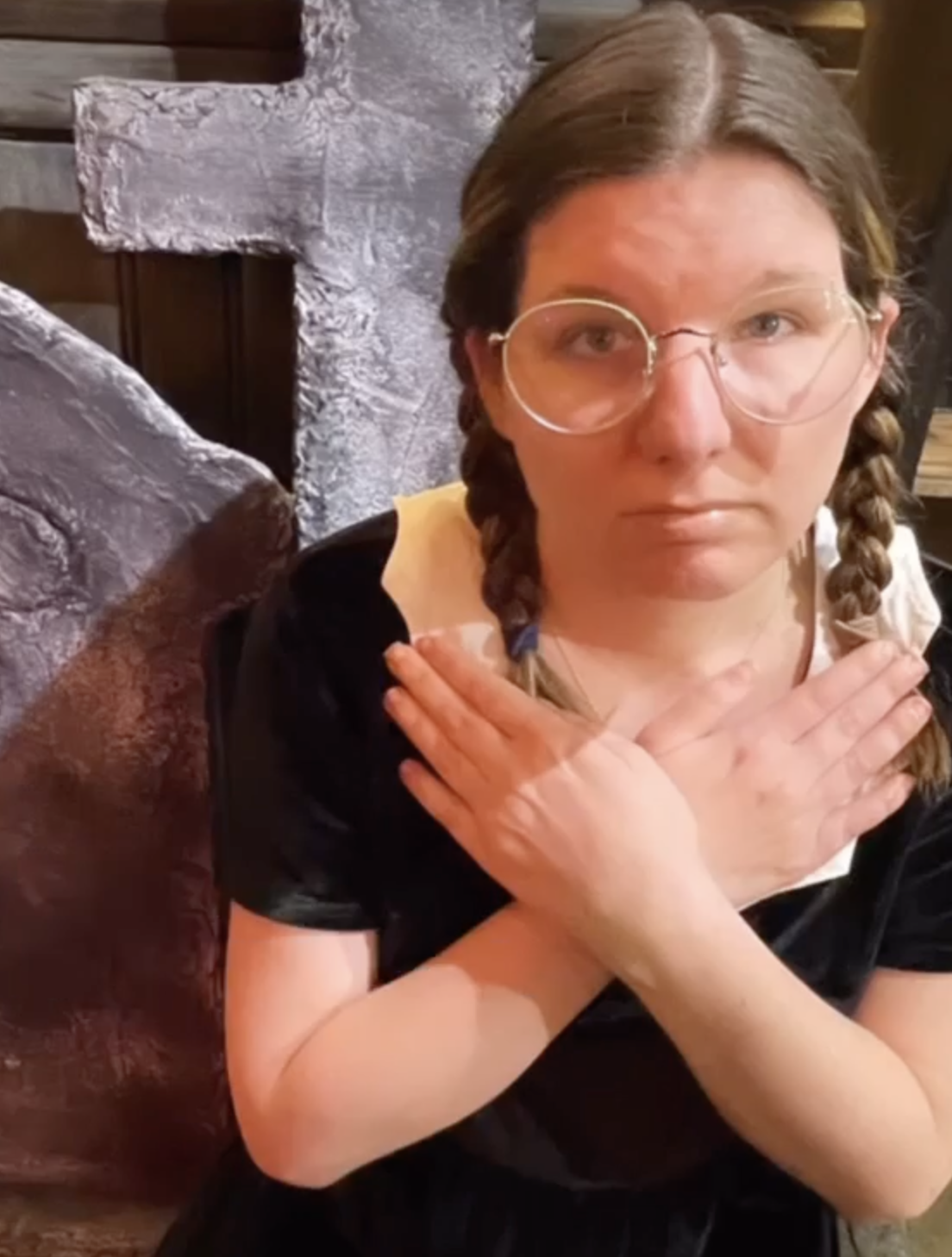 Amy, a young woman with braided hair and black dress stands in front of a crucifix with her hands folded across her chest.