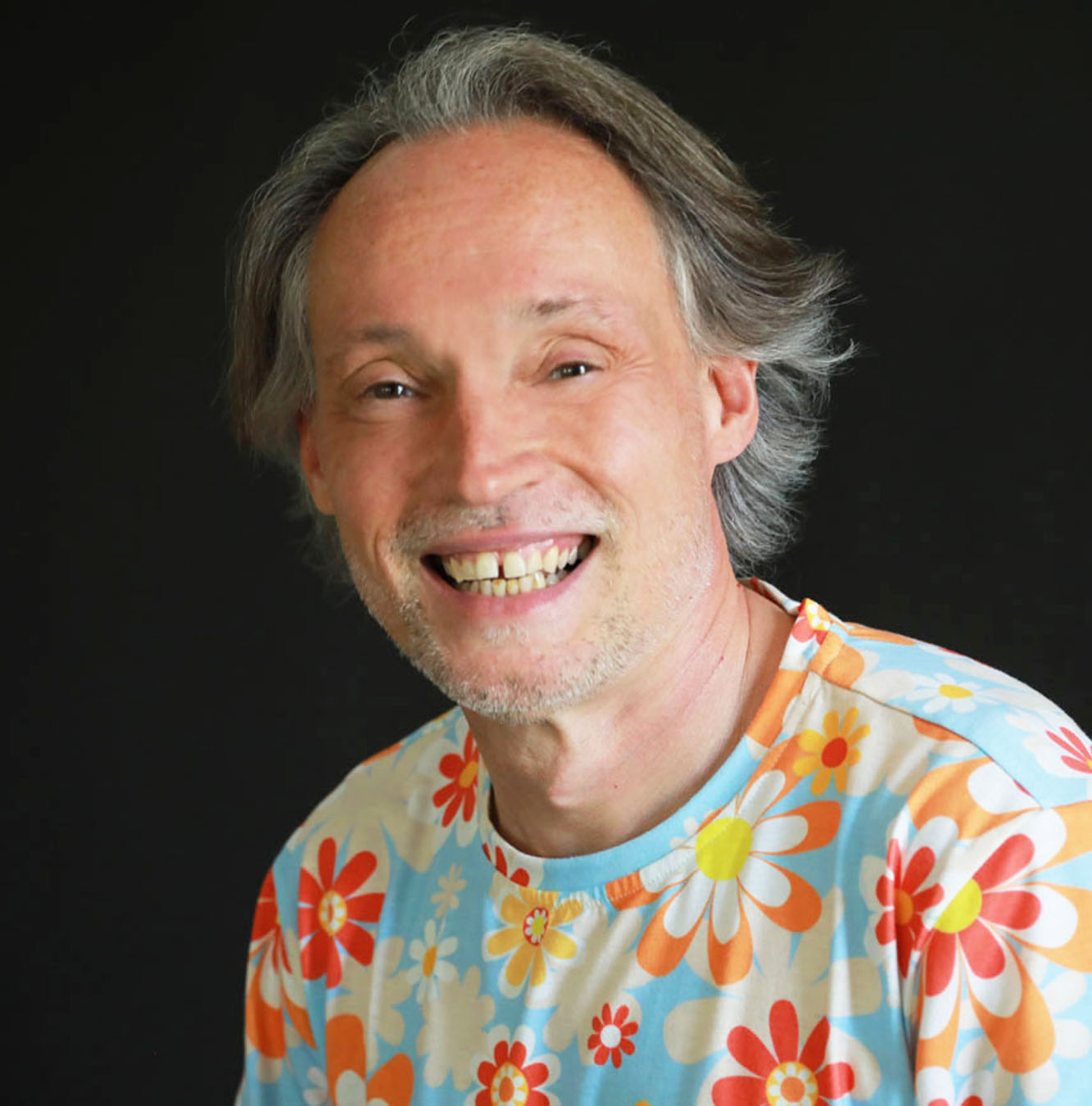 Man with long blong/ gray hair and big smile wearing a floral t-shirt.
