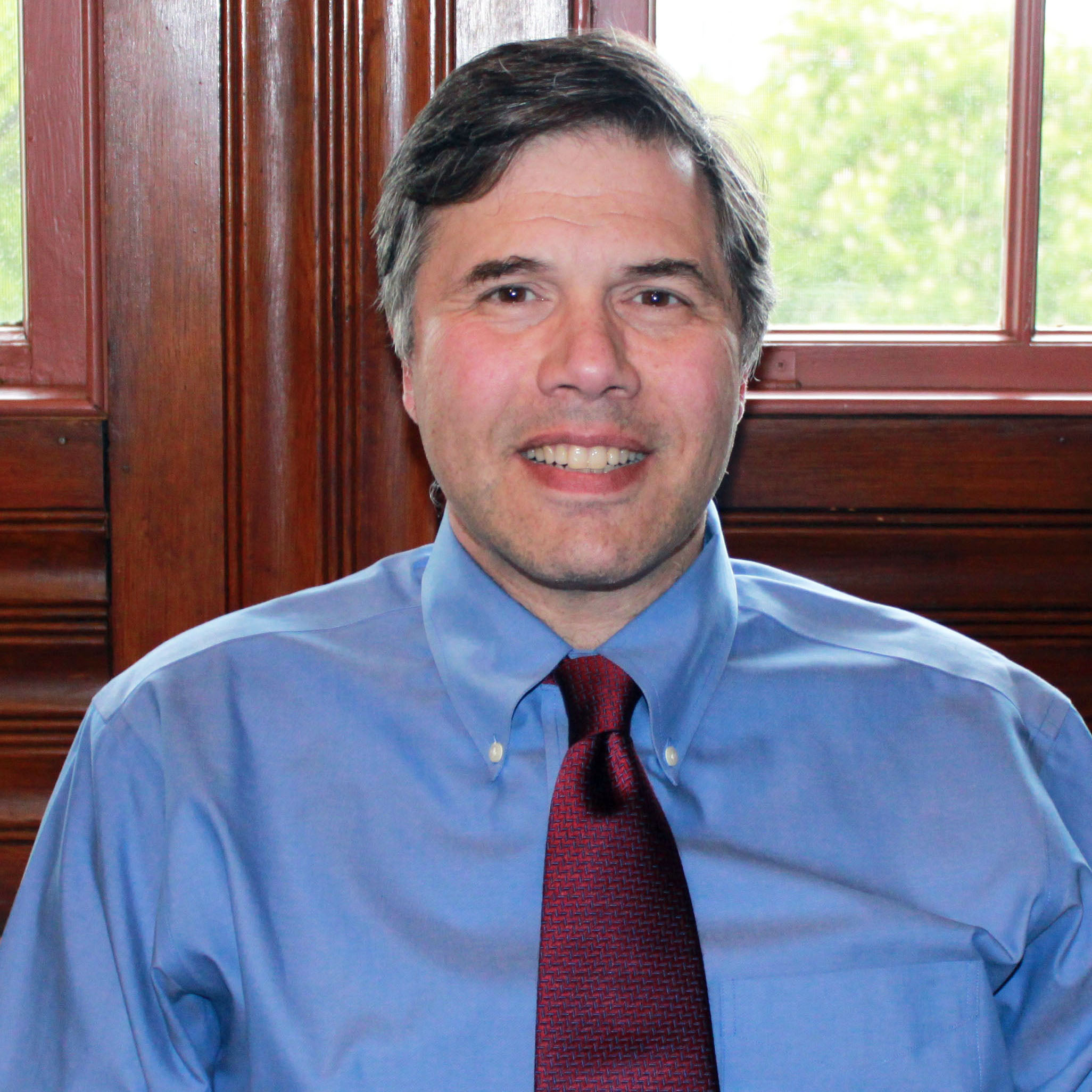 Man with graying hair in blue shirt and dark red tie in front of wood paneling and window.