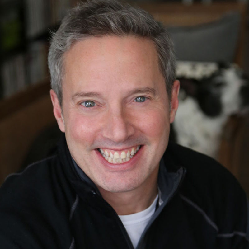 Light-skinned, grey-haired man wearing black, smiling.