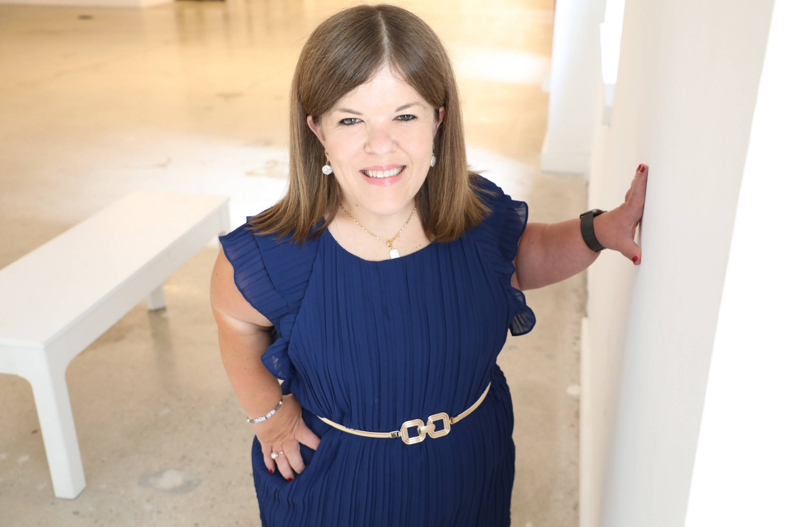 Woman smiling into the camera wearing a blue short sleeved dress with small gold belt.