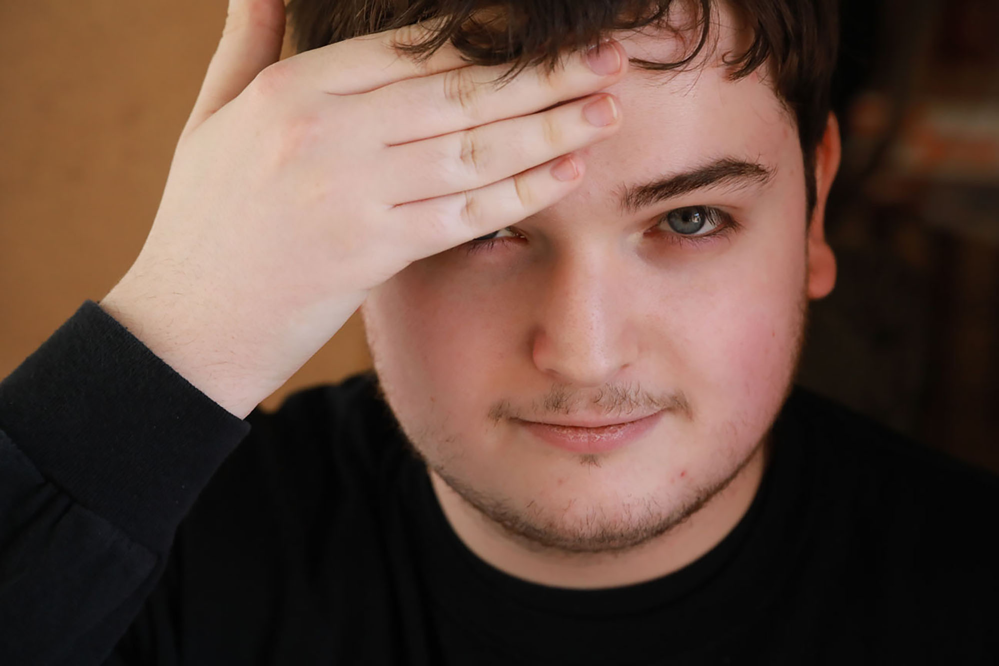 Man with brown hair looking into the lens and covering his right eye with his hand.
