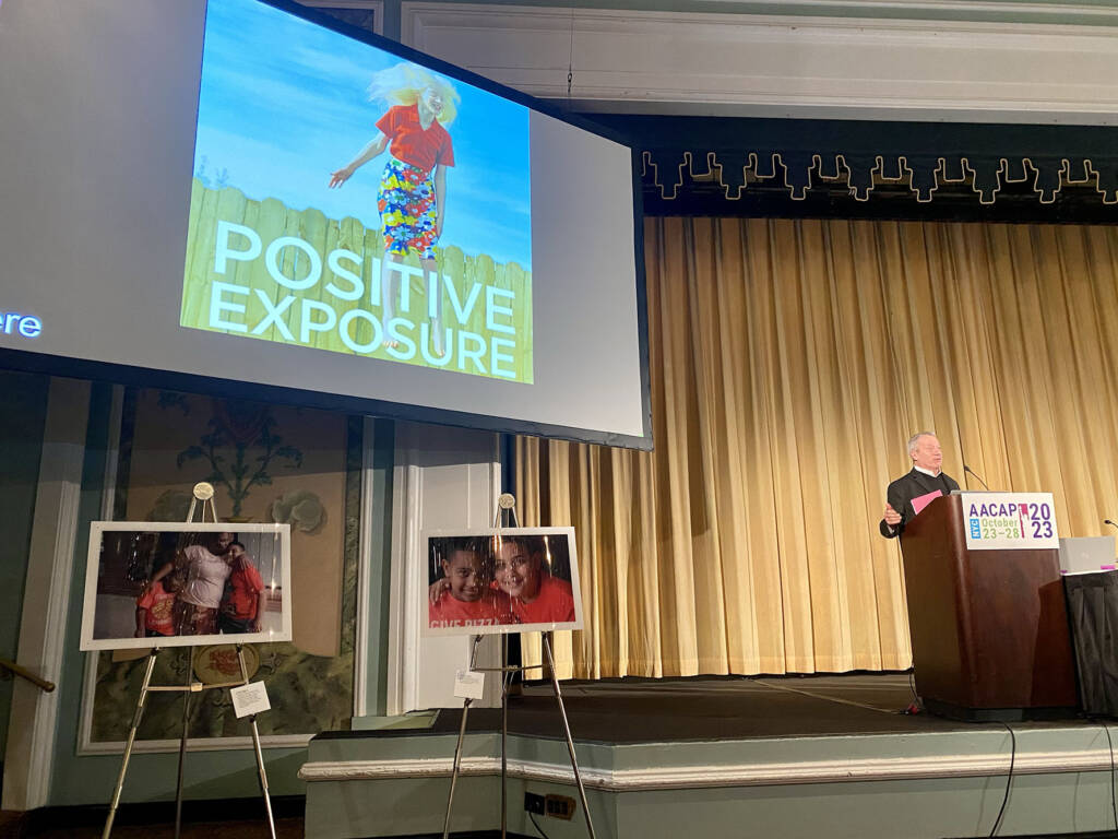 Rick at podium with Positive Exposure Logo on big screen.