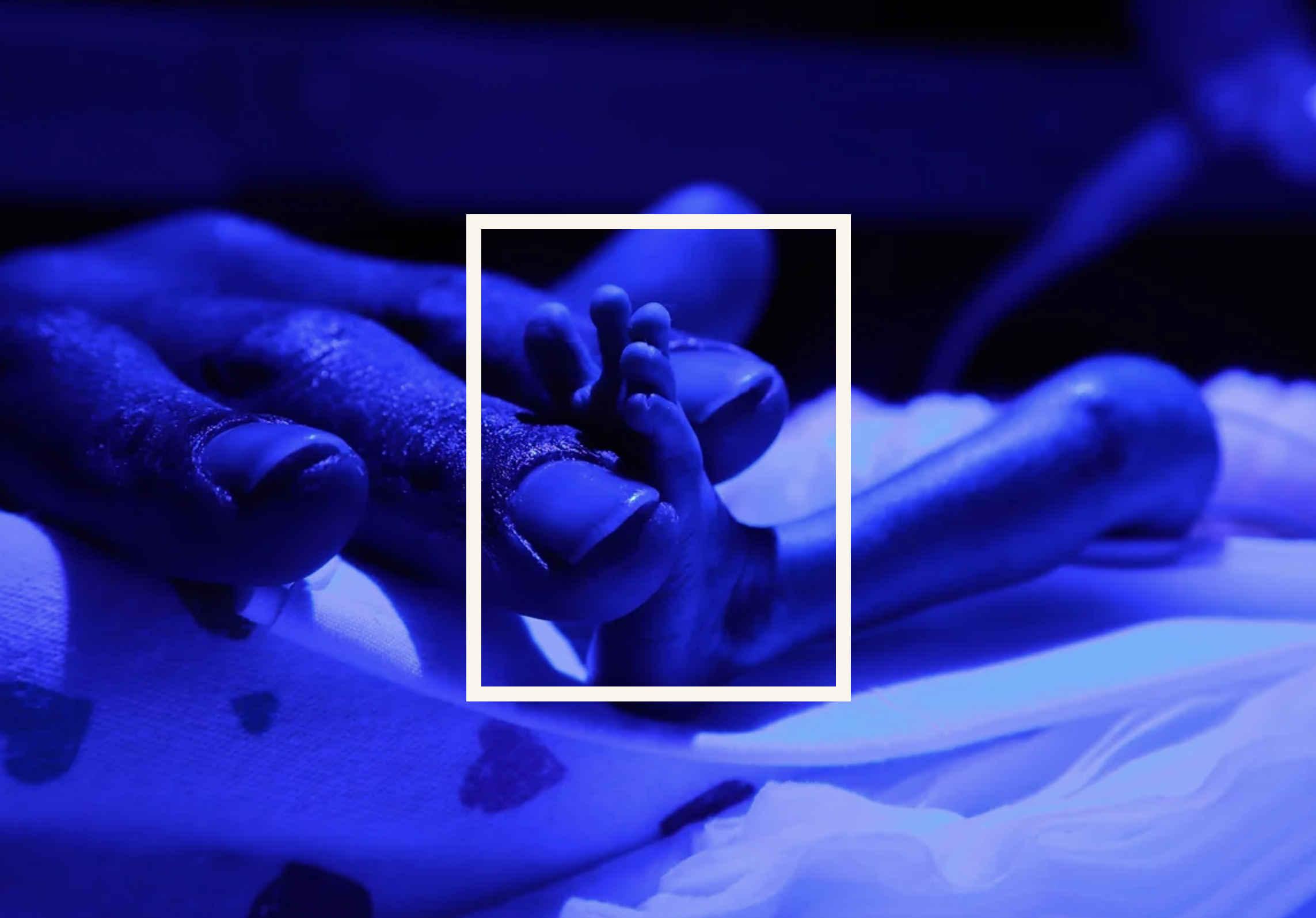 Image of man's fingers holding the tiny foot of an infant in the NICU. Image is bathed in blue light.
