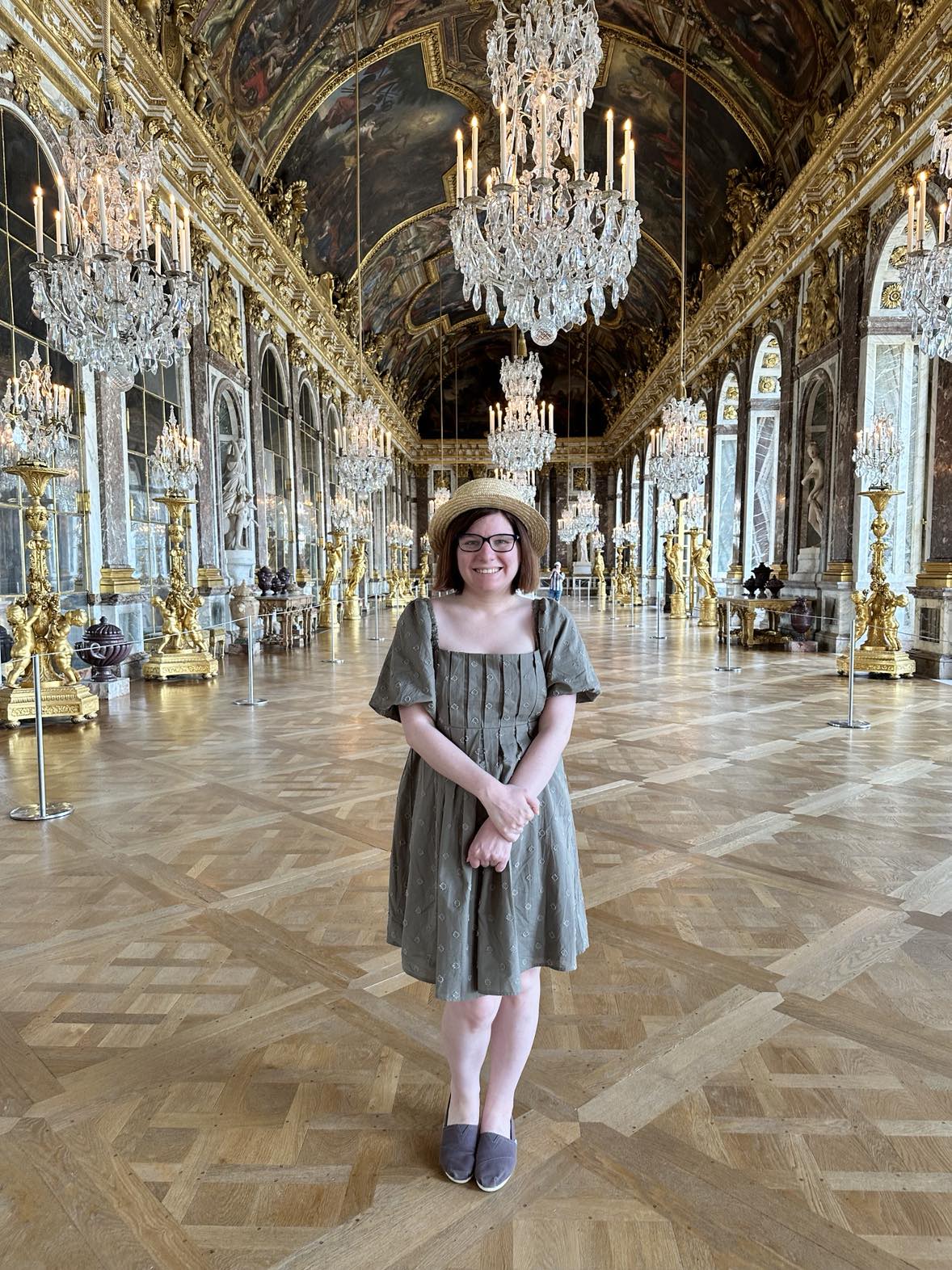 Amy, wearing a green dress, stands in a room in Versailles with enormous chandeliers.