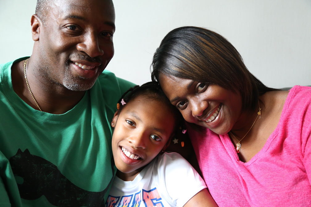 Young girl sitting between her mom and dad. All are smiling and looking into the camera lens.