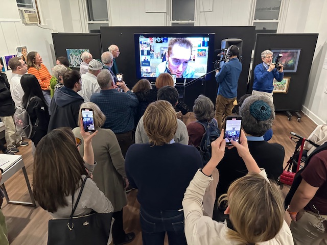 Group of people in the gallery gathering around artist who is on screen.