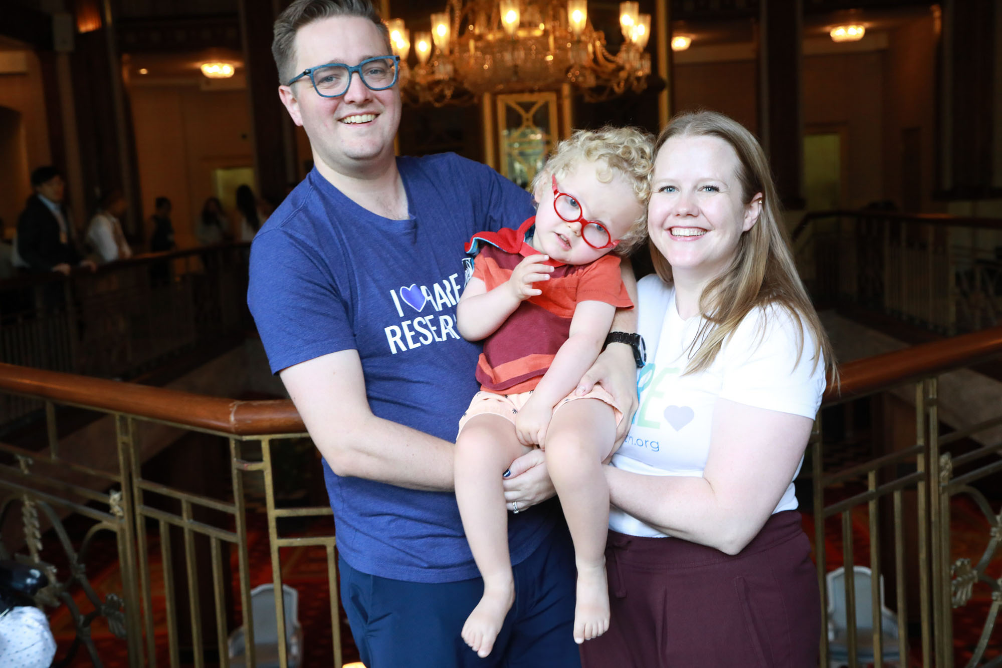 young girl wearing red glasses being held by both her parents, smiling