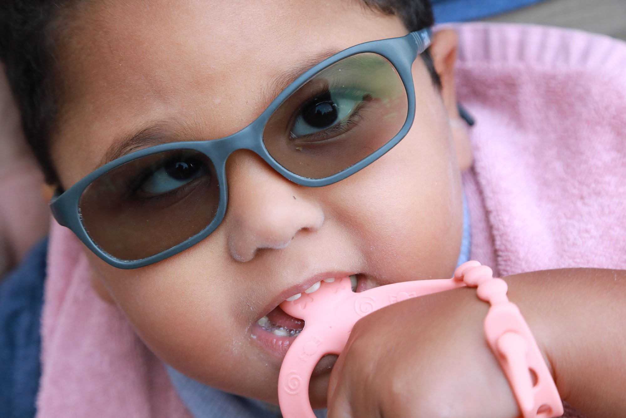 young girl with grey sunglasses and pink outfit looks into the camera while chewing on a pink toy