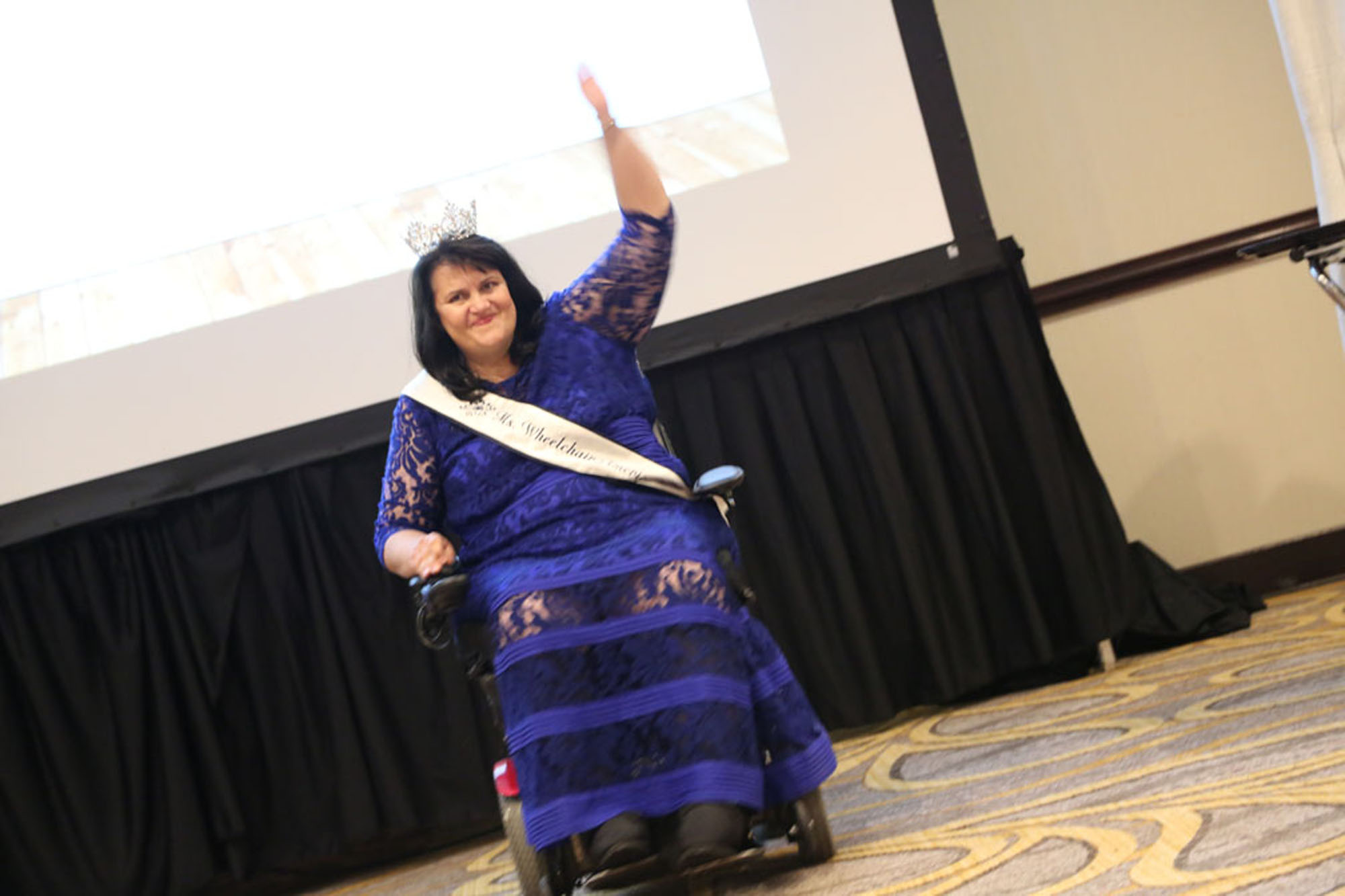 woman with klippel trenaunay syndrome wearing purple dress in a wheel chair, one arm raised