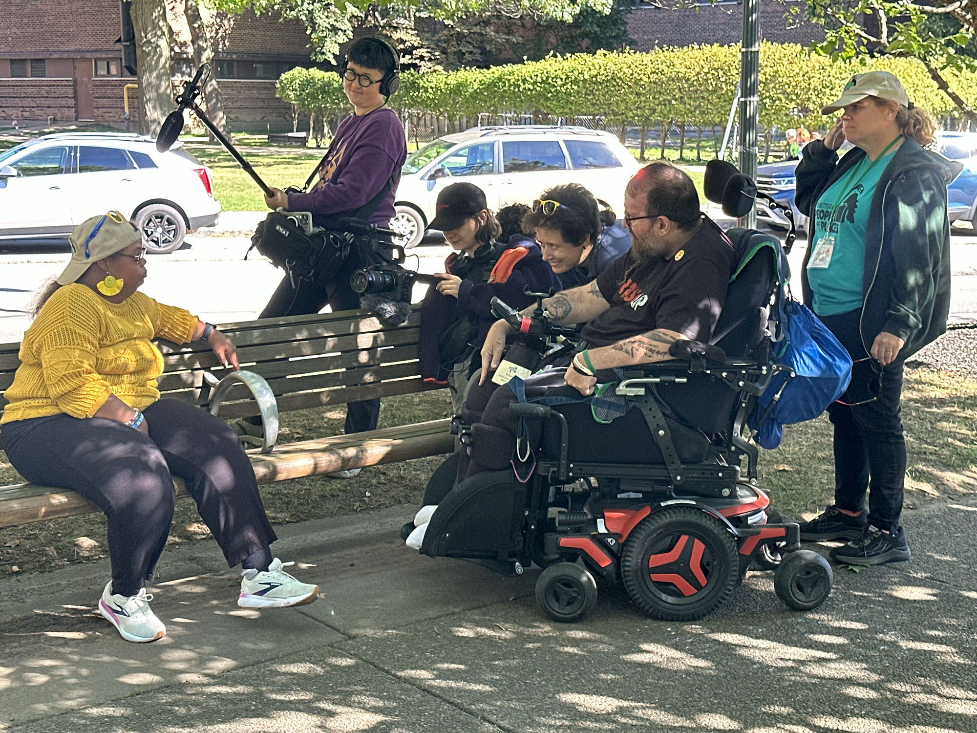 woman sitting on a park bench with a film crew, camera, mic on boom all focused on her. The interviewer is a man in a motorized Wheelchair.