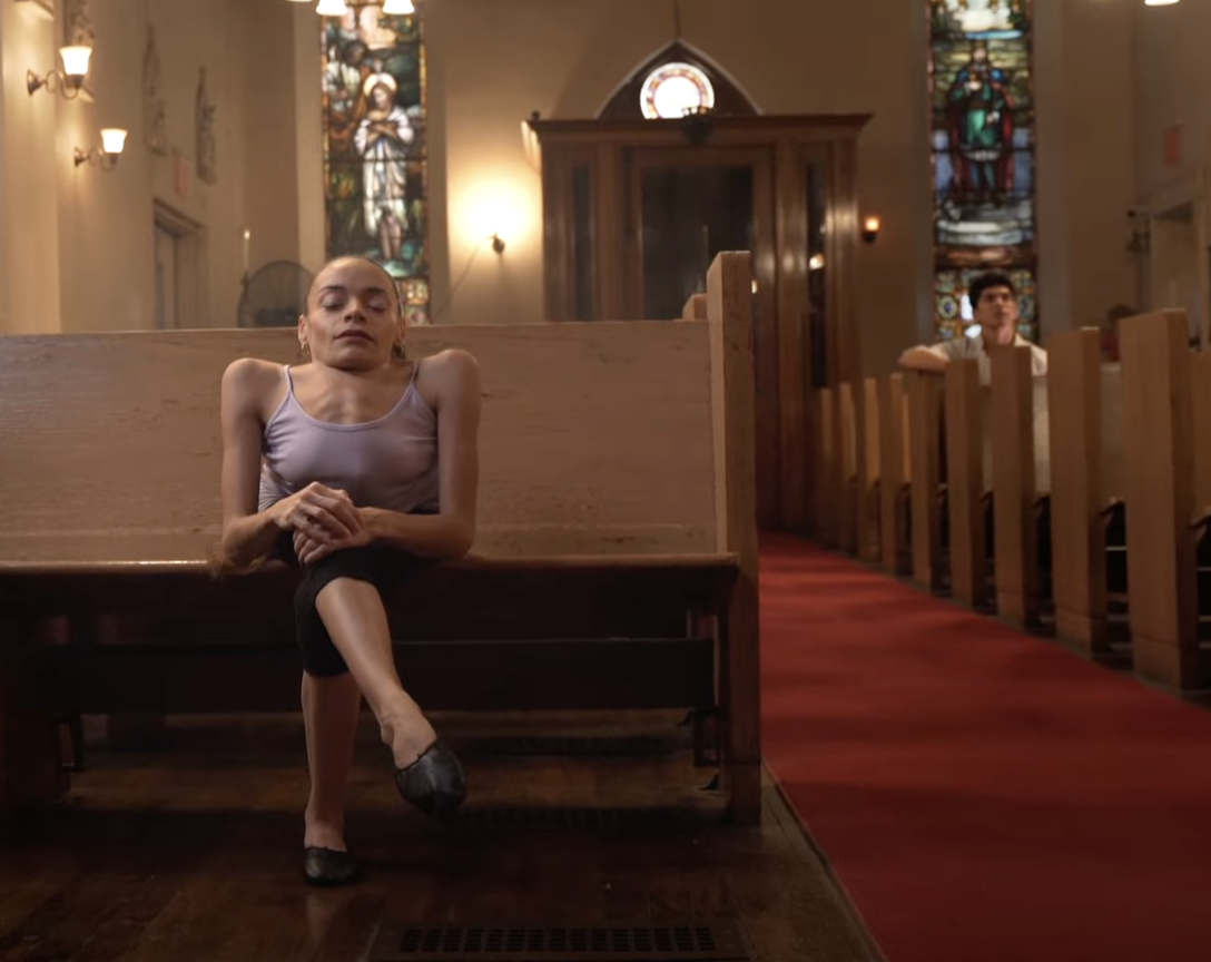 Image of a woman wearing a light blue tank tiop sitting in the front pew of a church. A man in a white shirt sits a few pews behind her on the right.