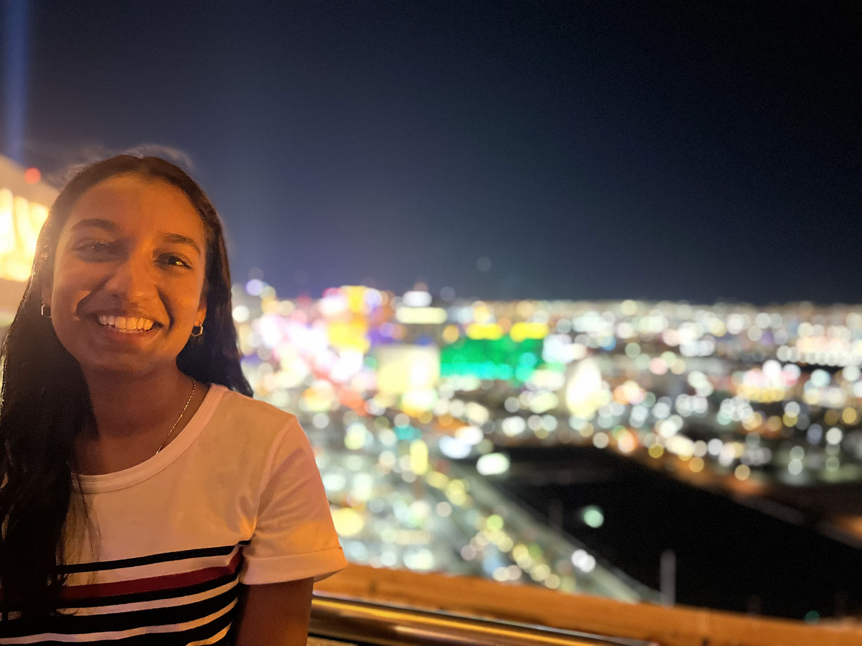 Woman with brown hair in white t shirt standing on balcony with cityscape and city lights in the background.