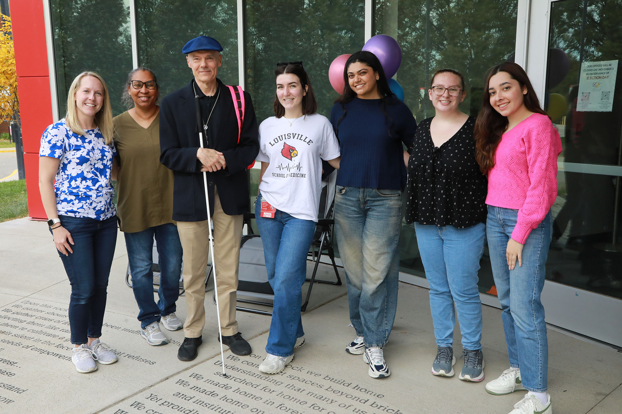 AX2A9741 Group of 8 people, 7 women in summer attire and man in dark blazer with blue beret holding a white cane and everyone is looking into the camera.