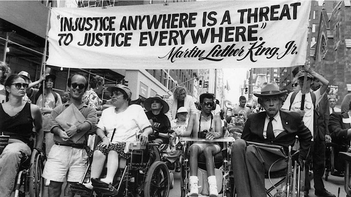Black and white image of several protesters in wheelchairs under a banner that reads Injustice anywhere is a threat to justice everywhere, Martin Luther King Jr.