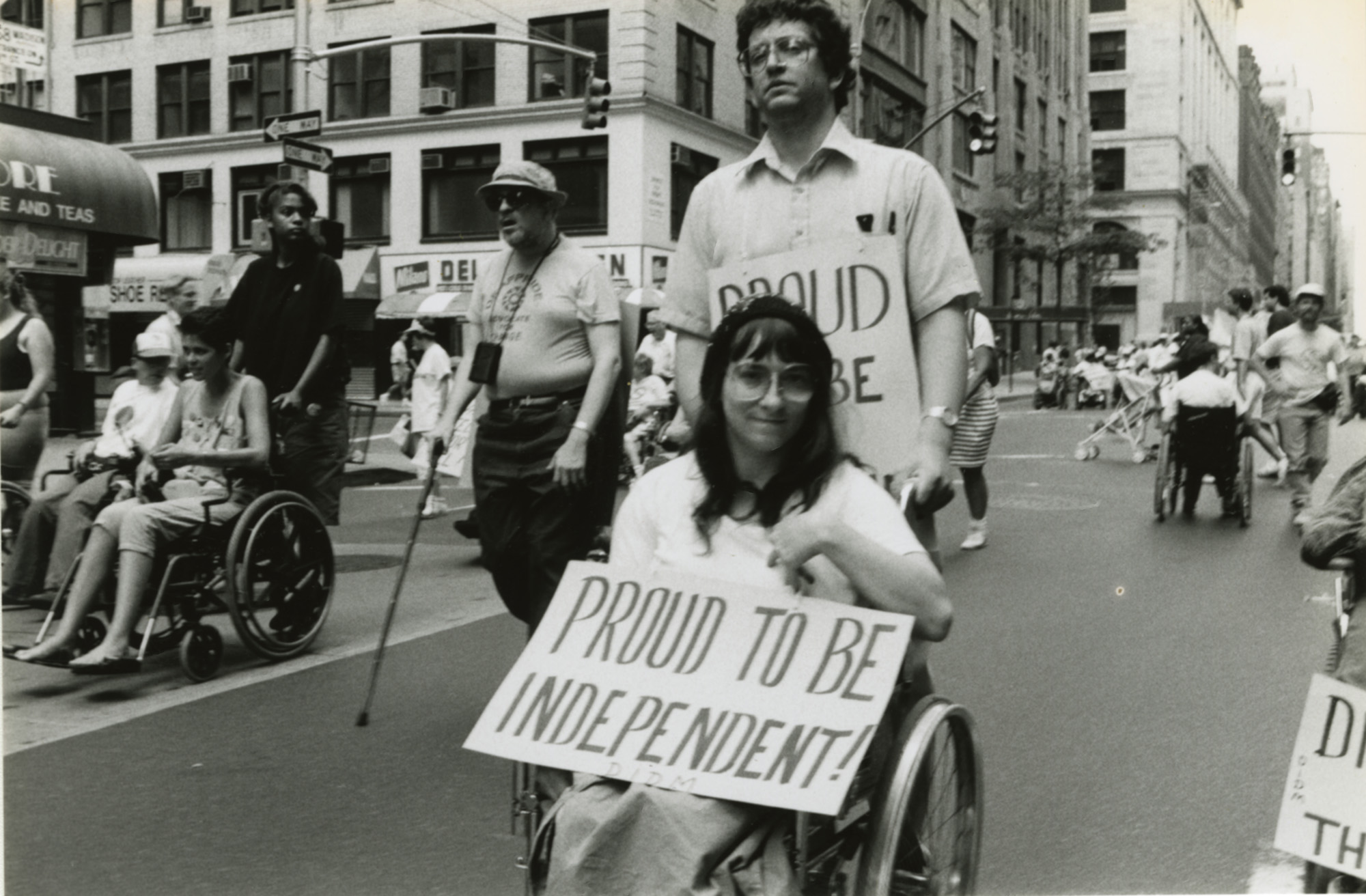A black and white photo of a woman in a wheelchair being pushed by a man also carrying a sign. The woman is holding a sign that reads, Proud to be independent.